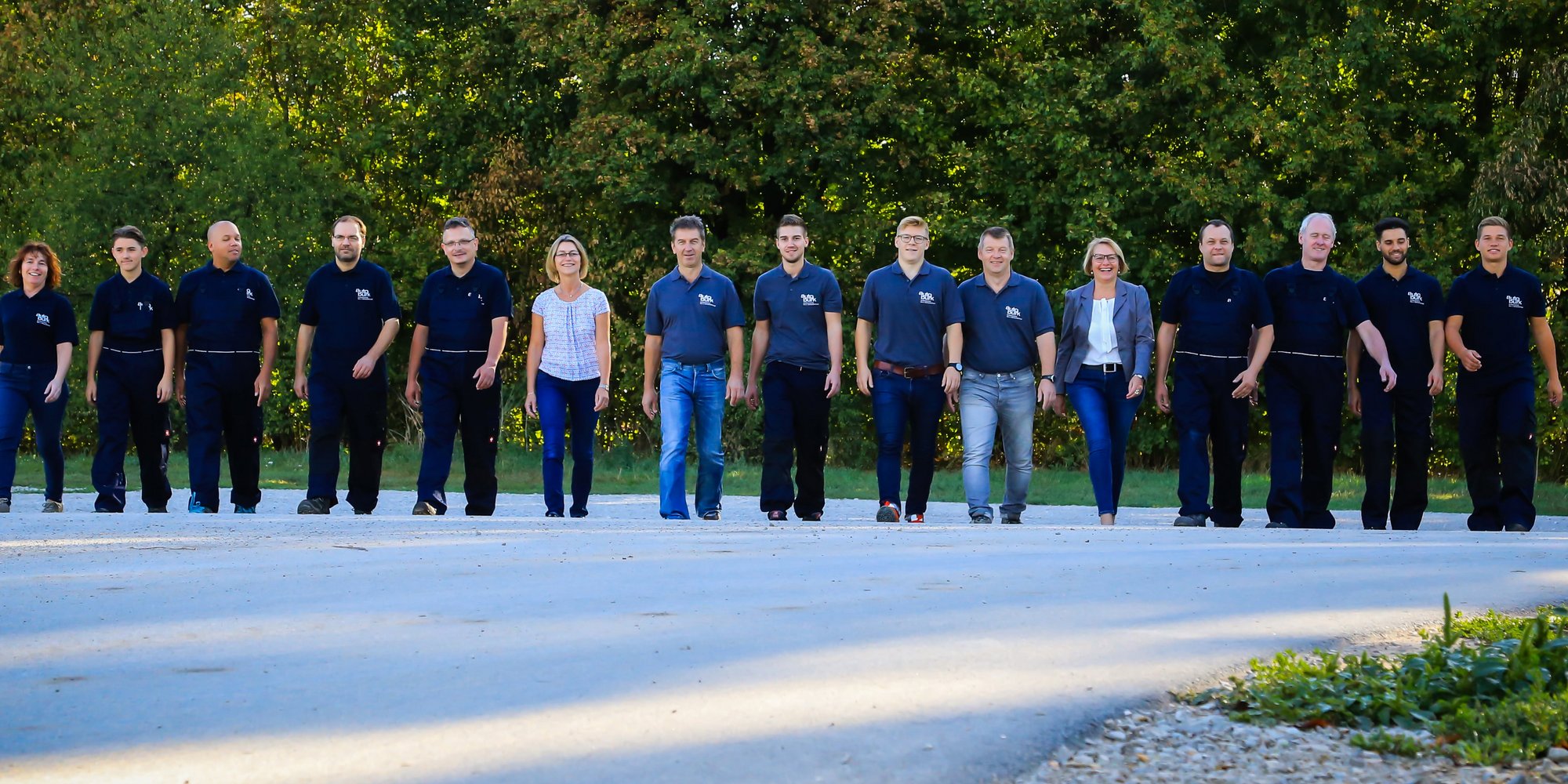 Eine Gruppe von Personen in Arbeitskleidung und läuft gemeinsam auf einer Straße, im Hintergrund Bäume.